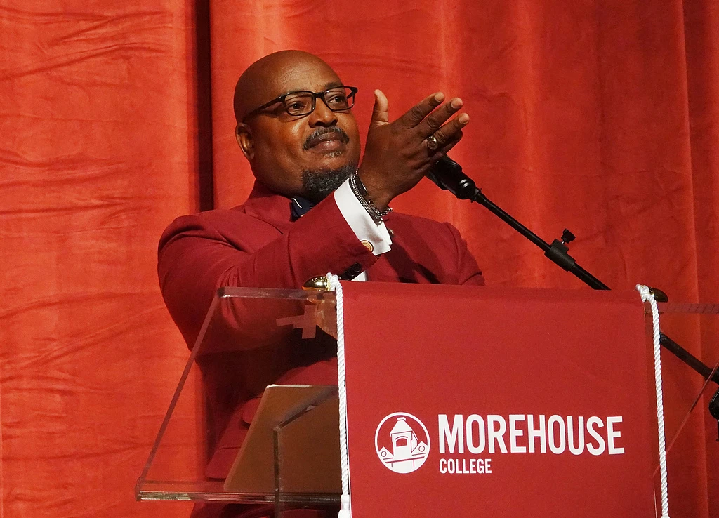 A man in a red suit is speaking at a podium with a Morehouse College banner on it, in front of a red curtain.