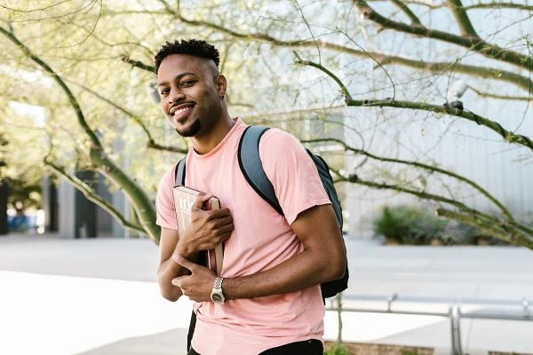 A man wearing a pink shirt and backpack holds a book, smiling outdoors with trees and a building in the background.